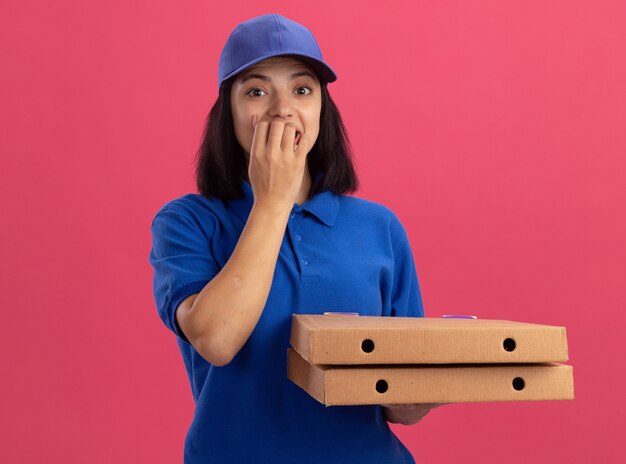 青いユニフォームとピザの箱を保持しているキャップの若い配達の女の子は、ピンクの壁の上に立っている彼女の爪を噛んでストレスと神経質になりました