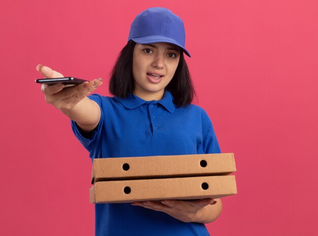 Giovane ragazza delle consegne in uniforme blu e cappuccio che tiene le scatole per pizza allungando il telefono cellulare in piedi sopra la parete rosa