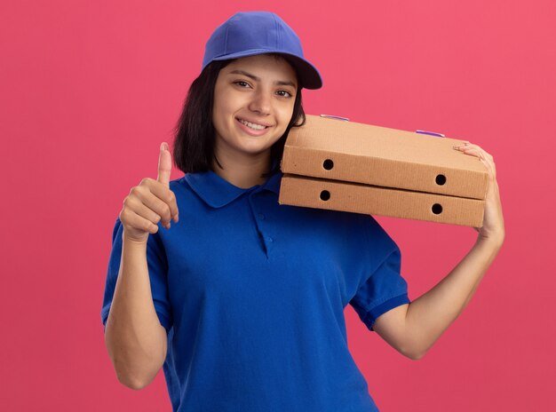 ピンクの壁の上に立って親指を示す幸せそうな顔でピザの箱を保持している青い制服と帽子の若い配達の女の子