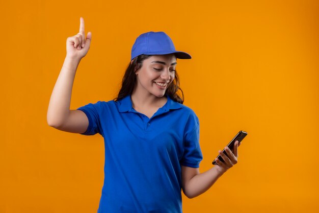 Молодая доставщица в синей форме и кепке держит смартфон в руке, глядя на него, указывая пальцем вверх, имея отличную идею, уверенно улыбаясь, стоя на желтом фоне