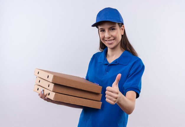 Young delivery girl in blue uniform and cap holding stack of pizza boxes looking at camera with confident smile on face showing thumbs up 