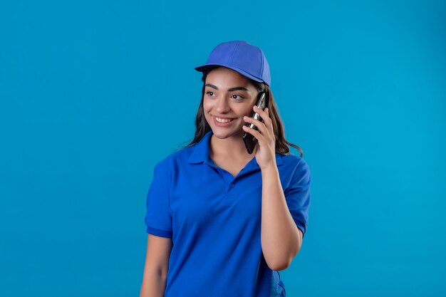 파란색 제복을 입은 젊은 배달 소녀와 모자는 파란색 배경 위에 서있는 휴대 전화에 친절한 이야기를 제쳐두고 웃고 있습니다.