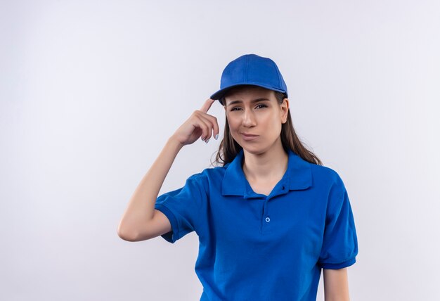 Young delivery girl in blue uniform and cap looking at camera confused pointing her temple for mistake, forgot 