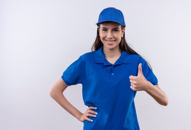 Молодая доставщица в синей униформе и кепке смотрит в камеру, весело улыбаясь, показывает палец вверх