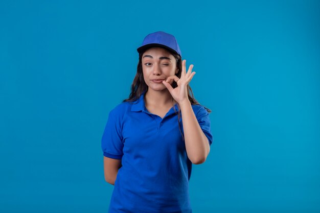 Молодая доставщица в синей униформе и кепке смотрит в камеру, подмигивая, делая жест тишины, как будто закрывая рот застежкой-молнией, стоящей на синем фоне