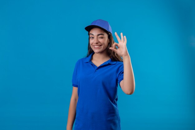 Young delivery girl in blue uniform and cap looking at camera winking smiling cheerfully doing ok sign standing over blue background