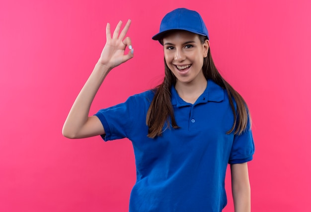 Foto gratuita giovane ragazza di consegna in uniforme blu e berretto guardando camra sorridendo allegramente mostrando segno ok