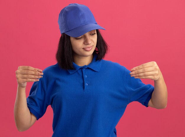 青い制服を着た若い配達の女の子と指で自信を持って身振りで示すキャップ、ピンクの壁の上に立っているボディーランゲージの概念