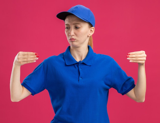 Foto gratuita giovane ragazza delle consegne in uniforme blu e berretto dall'aria sicura che gesticola con le mani come tenere qualcosa in piedi sul muro rosa