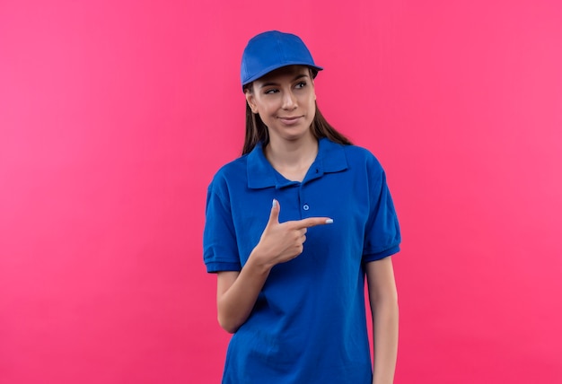 Молодая доставщица в синей форме и кепке выглядит уверенно с улыбкой на лице, указывая пальцем на бок