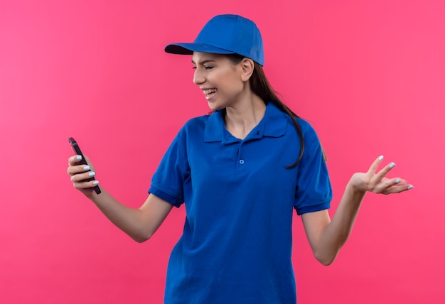 Foto gratuita giovane ragazza delle consegne in uniforme blu e cappuccio guardando lo schermo dello smartphone deluso