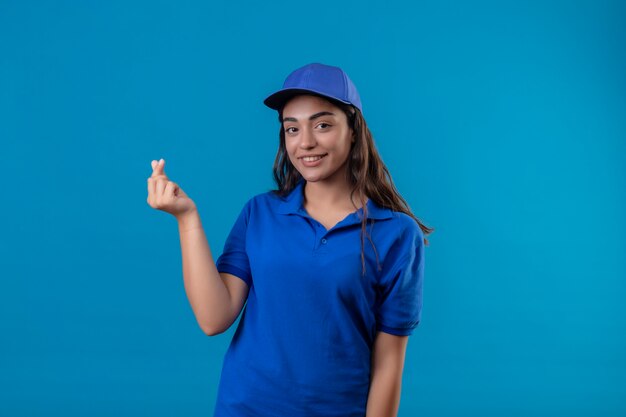 Молодая доставщица в синей форме и кепке делает денежный жест, уверенно улыбаясь, глядя в камеру, стоящую на синем фоне