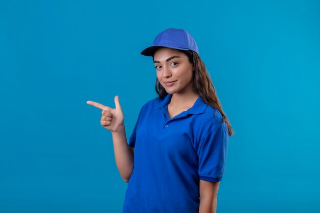 Молодая доставщица в синей униформе и кепке, указывая пальцем в сторону, улыбается уверенно, счастливо и позитивно, стоя на синем фоне
