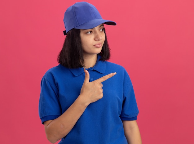 Young delivery girl in blue uniform and cap pointing with index figner to the side looking confident standing over pink wall