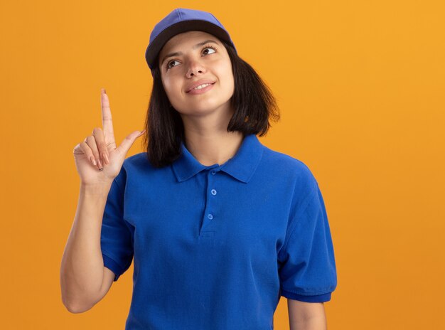 Young delivery girl in blue uniform and cap pointing with index figner up smiling standing over orange wall