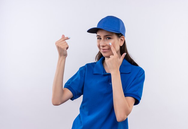 Молодая доставщица в синей униформе и кепке трет пальцы, указывая пальцем в глаза, ожидая оплаты