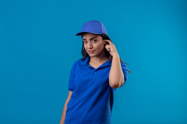 Young delivery girl in blue uniform and cap scratching head looking at camera with confuse expression on face having doubts standing over blue background