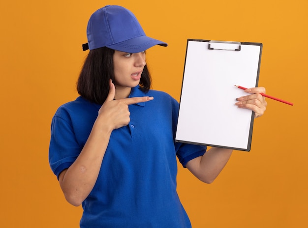 Young delivery girl in blue uniform and cap showing clipboard with blank pages and pencil pointing with index finger at them confused standing over orange wall