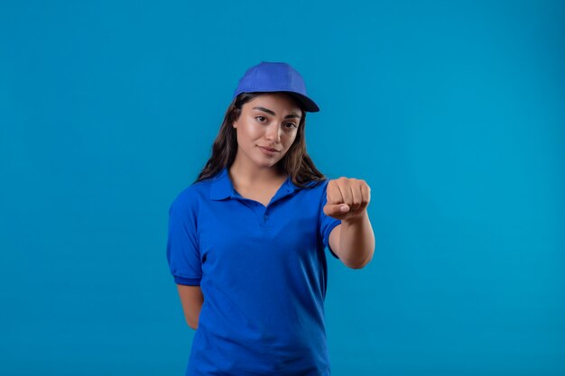 Молодая доставщица в синей форме и кепке показывает кулак в камеру с серьезным уверенным выражением лица, стоящим на синем фоне