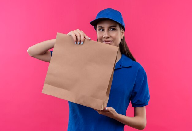 Young delivery girl in blue uniform and cap showing paper package looking aside smiling friendly 