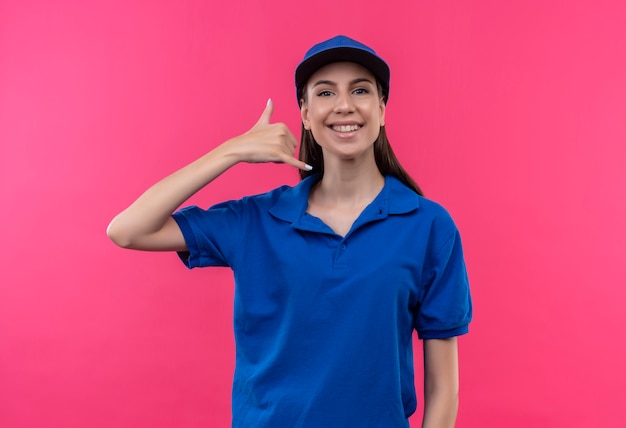 Young delivery girl in blue uniform and cap smiling cheerfully making call me gesture 