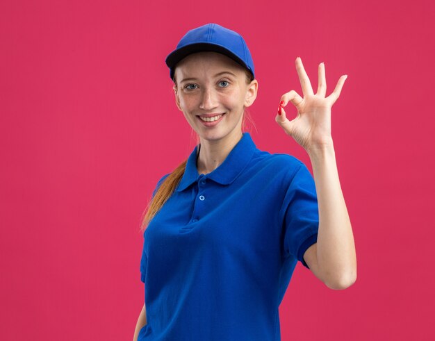 Молодая доставщица в синей форме и кепке, уверенно улыбаясь, делает хорошо, знак стоит над розовой стеной