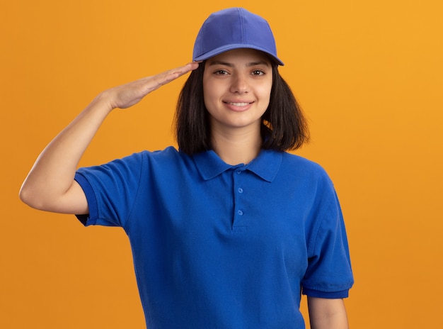 Young delivery girl in blue uniform and cap  smiling confident saluting standing over orange wall