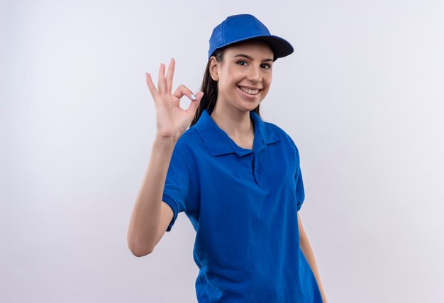 Молодая доставщица в синей форме и кепке уверенно улыбается, показывая знак ОК