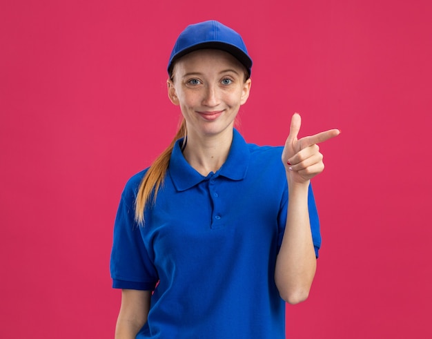 Молодая доставщица в синей форме и кепке дружелюбно улыбается, указывая указательным пальцем в сторону, стоя над розовой стеной