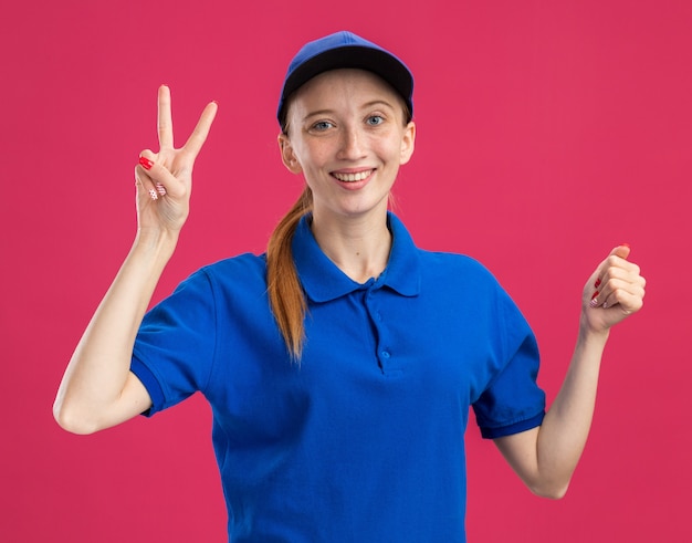Молодая доставщица в синей форме и кепке дружелюбно улыбается, показывая знак v и сжатый кулак, стоит над розовой стеной