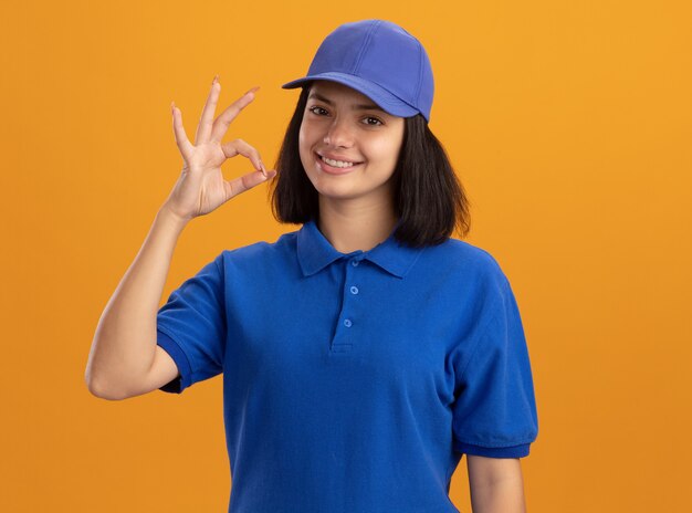파란색 유니폼과 모자에 젊은 배달 소녀 오렌지 벽 위에 서있는 확인 서명을 보여주는 웃 고
