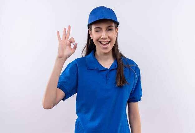 Молодая доставщица в синей униформе и кепке улыбается и уверенно подмигивает, показывая знак ОК