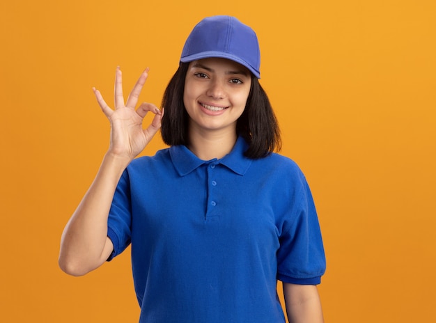 Foto gratuita giovane ragazza di consegna in uniforme blu e cappuccio sniling che mostra segno giusto in piedi sopra la parete arancione