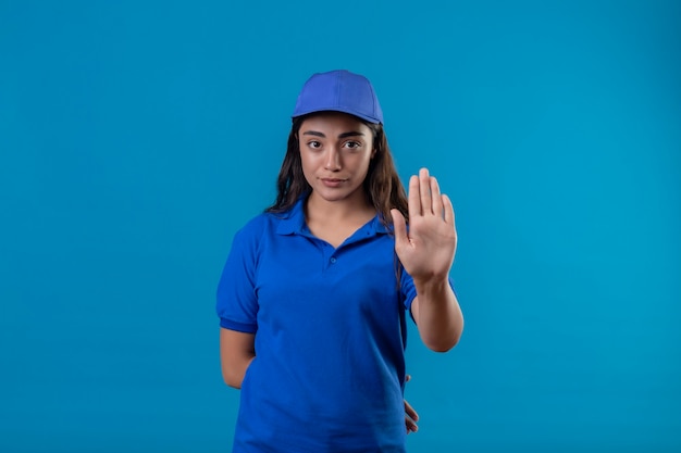 Free photo young delivery girl in blue uniform and cap standing with open hand doing stop sign with serious and confident expression defense gesture over blue background