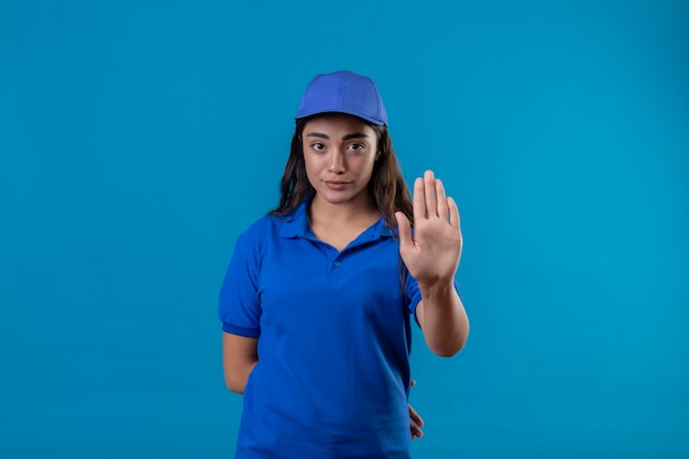 Молодая доставщица в синей форме и кепке, стоящая с открытой рукой, делает знак остановки с серьезным и уверенным выражением защитного жеста на синем фоне