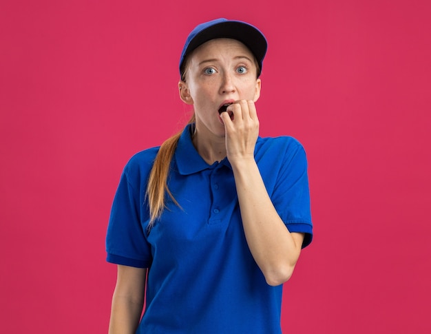 Giovane ragazza delle consegne in uniforme blu e berretto stressato e nervoso che si morde le unghie in piedi sul muro rosa