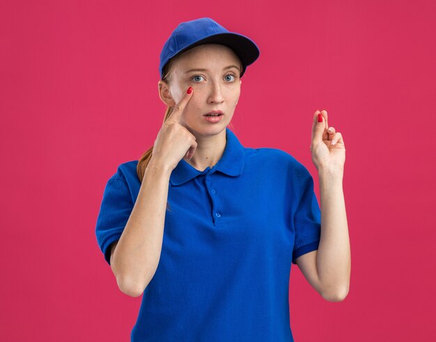 Giovane ragazza delle consegne in uniforme blu e berretto sorpreso indicando con il dito indice l'occhio