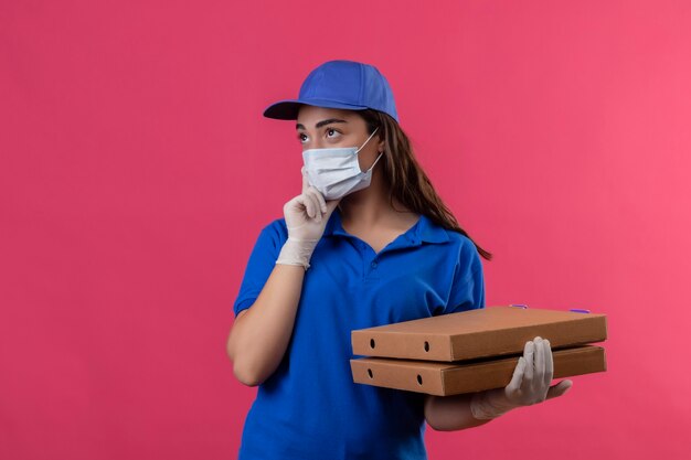 Молодая доставщица в синей униформе и кепке в защитной маске и перчатках держит коробки для пиццы, глядя в сторону с задумчивым выражением лица, думая, стоя на розовом фоне