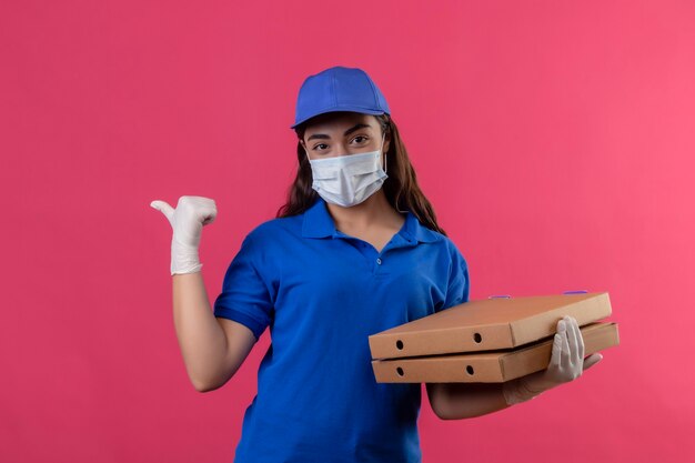 Young delivery girl in blue uniform and cap wearing facial protective mask and gloves holding pizza boxes looking at camera smiling confident positive and happy pointing with thumb to the side