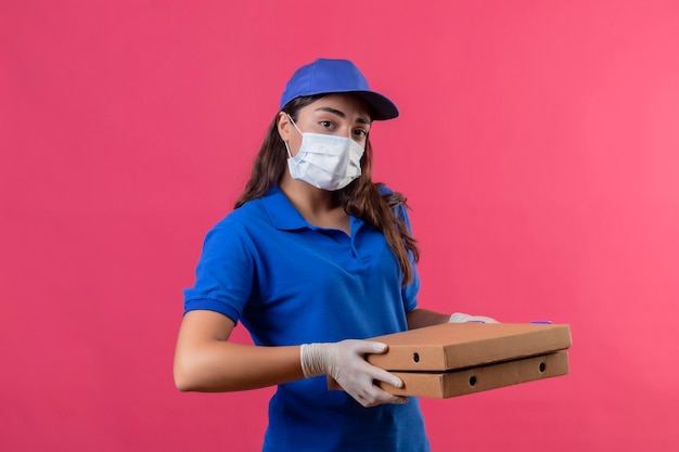 Young delivery girl in blue uniform and cap wearing facial protective mask and gloves holding pizza boxes looking at camera with serious confident facial expression standing over pink backgroun