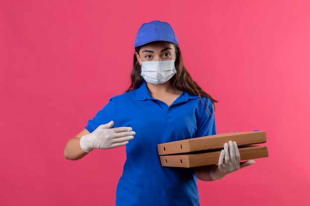 Young delivery girl in blue uniform and cap wearing facial protective mask and gloves holding pizza boxes pointing with arm of hand looking confident standing over pink background