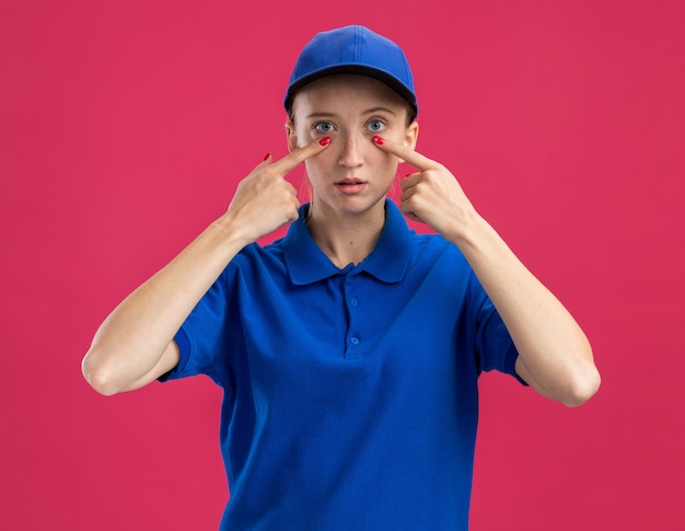 Giovane ragazza delle consegne in uniforme blu e berretto con una faccia seria che punta con il dito indice agli occhi in piedi sul muro rosa