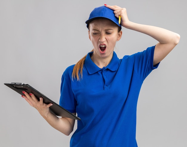 무료 사진 파란색 제복을 입은 젊은 배달 소녀와 클립 보드를 들고 모자가 흰 벽 위에 서있는 실수로 그녀의 머리에 손으로 혼동