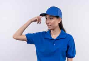 Бесплатное фото Молодая доставщица в синей униформе и кепке выглядит сбитой с толку, указывая висок на ошибку
