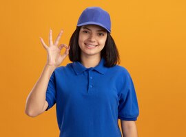 Молодая доставщица в синей форме и кепке с улыбкой показывает знак ок, стоящий над оранжевой стеной