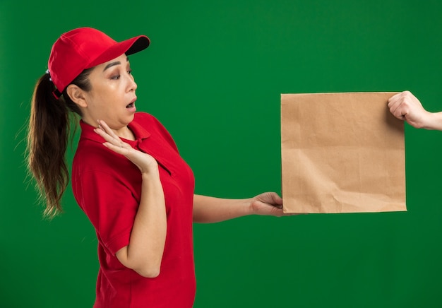 Молодая доставщица в красной форме и кепке отказывается получать бумажный пакет, глядя на него с удивлением, стоя у зеленой стены