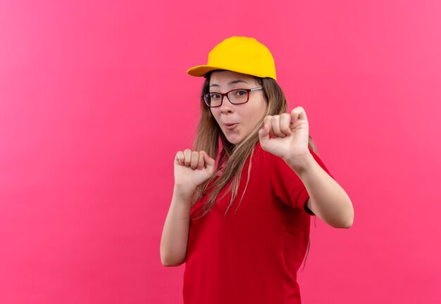 Young delivery girl in red polo shirt and yellow cap exited and happy clenching fists dancing 