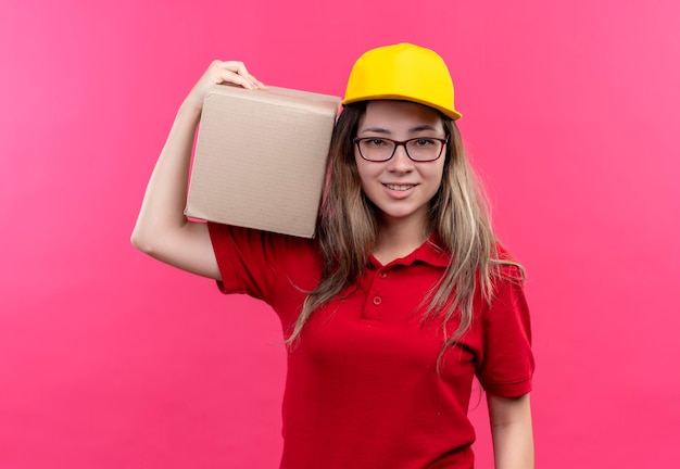 Young delivery girl in red polo shirt and yellow cap holding box package looking at camera smiling confident 