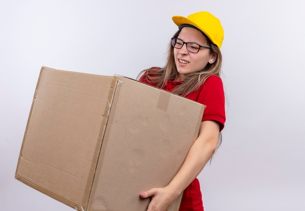 Young delivery girl in red polo shirt and yellow cap holding box package looking unwell suffering from weight of box 