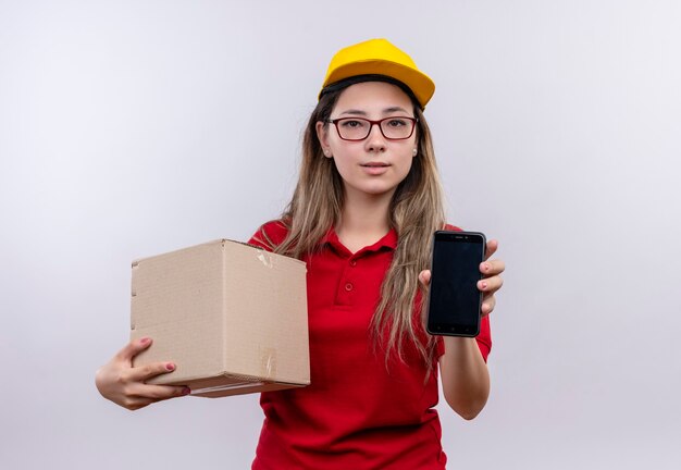 Young delivery girl in red polo shirt and yellow cap holding box package showing smartphone looking at camera with serious confidentexpression on face 